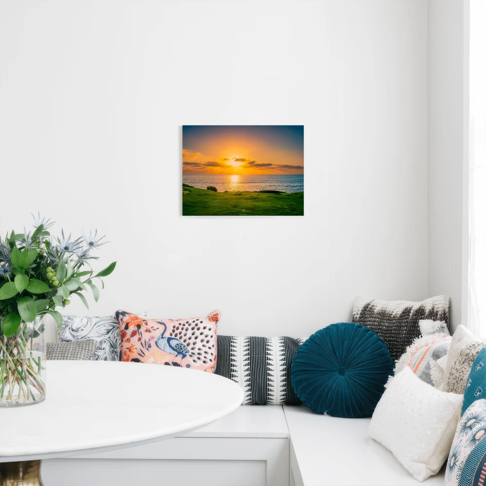 A rectangular wall art print showing a sunset over a grassy shoreline, displayed above a white bench with patterned pillows in a bright modern room.