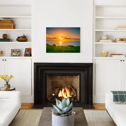 A square canvas wall art print depicting a vibrant sunset over a grassy field and ocean, displayed in a modern living room setting above a black fireplace.