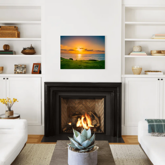 A square canvas wall art print depicting a vibrant sunset over a grassy field and ocean, displayed in a modern living room setting above a black fireplace.
