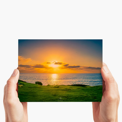 A person holds a rectangular art print or poster showing a coastal sunset scene with green grass in the foreground, blue water, and an orange-yellow sky, against a plain light background.