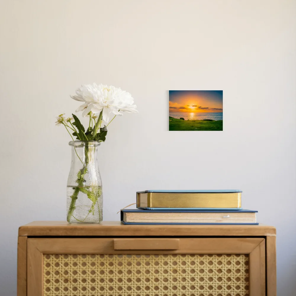 A small square wall art print showing a sunset over water with green grass in the foreground, displayed on a light wall above a wooden cabinet with a vase of white flowers and stacked books.