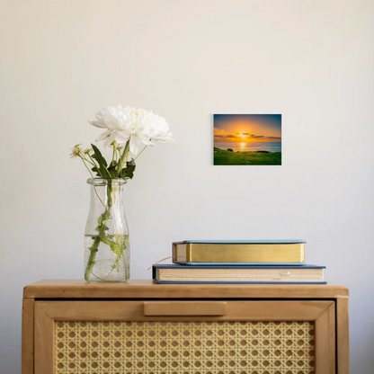 A small square wall art print showing a sunset over water with green grass in the foreground, displayed on a light wall above a wooden cabinet with a vase of white flowers and stacked books.